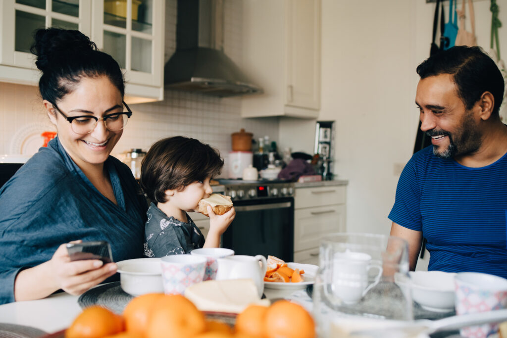 A mother glances at her cell phone while carrying a young boy, eating bread, on her lap. The father looks on, smiling at his son.