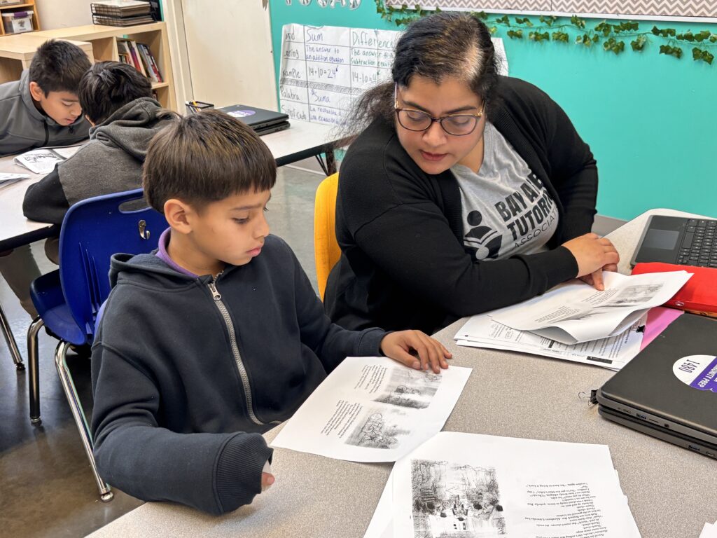 An adult woman sits beside an elementary school boy and they look at printed packets with primary source documents. They sit in a classroom together.