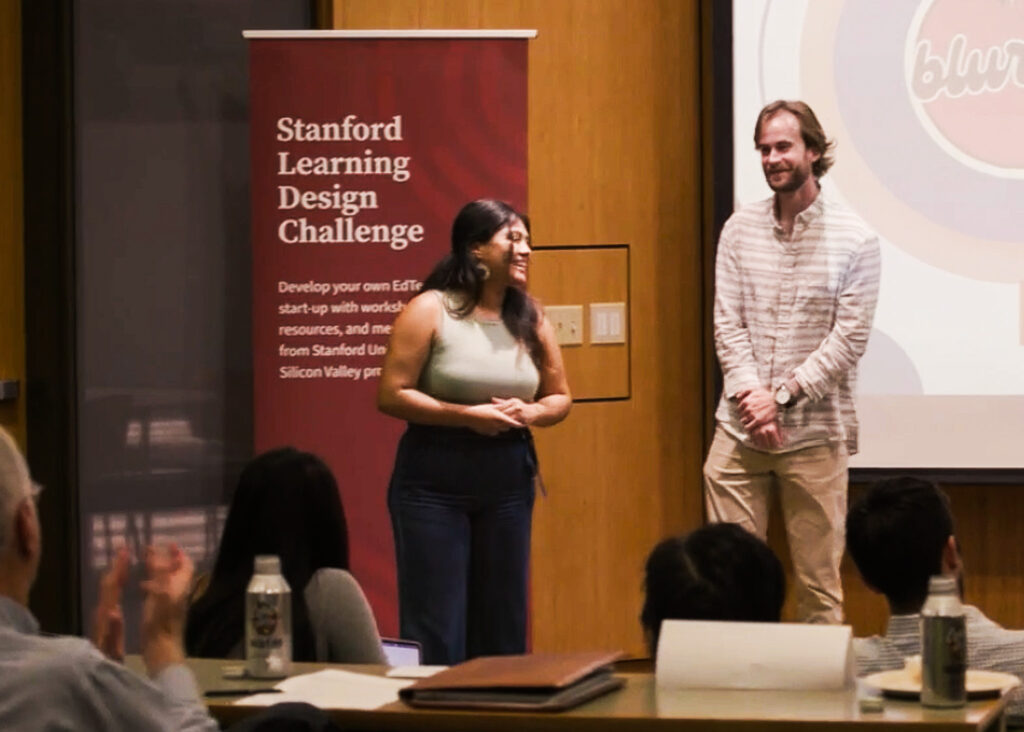 Two graduate students stand, smiling, in front of a table of clapping judges. There is a sign that says Stanford Learning Design Challenge.
