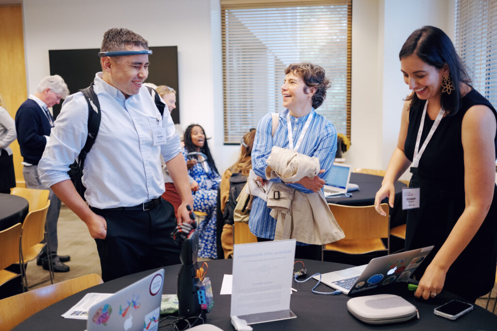 Three people stand at a table, laughing. One of them is wearing a neural sensor headband, and another looks at a laptop computer.