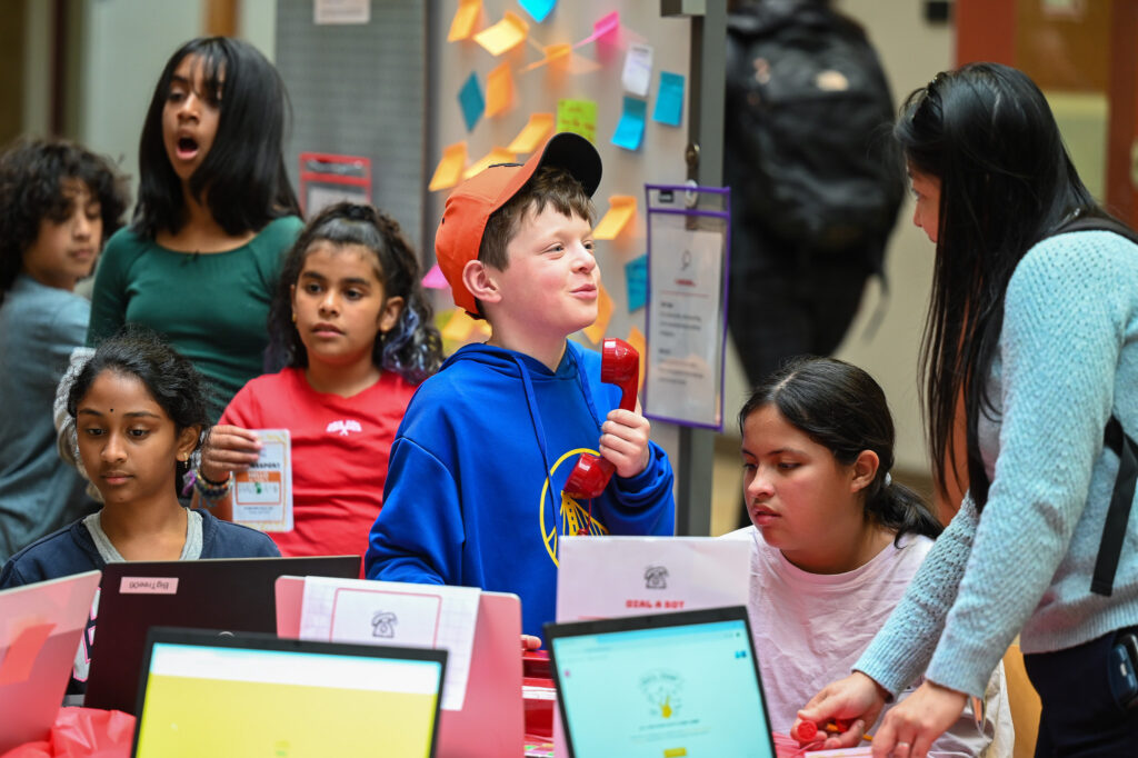 A young boy holding a red telephone handset, surrounded by his classmates who look at laptops, asks an adult woman a question.