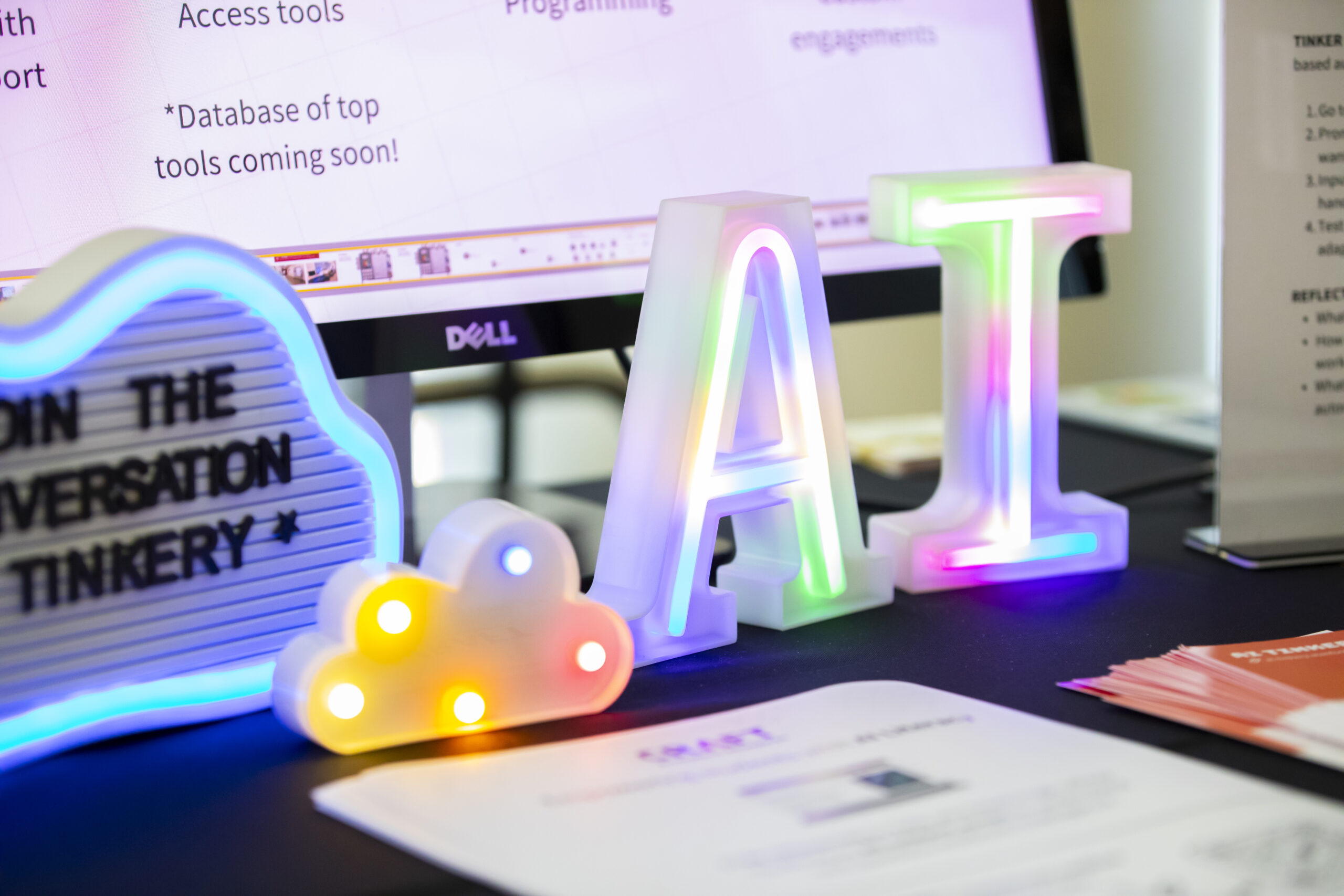 A closeup of a table set up with a desktop computer, glowing plastic decorative letters that say "AI", and a small board with letters that say, "Join the conversation, AI Tinkery."