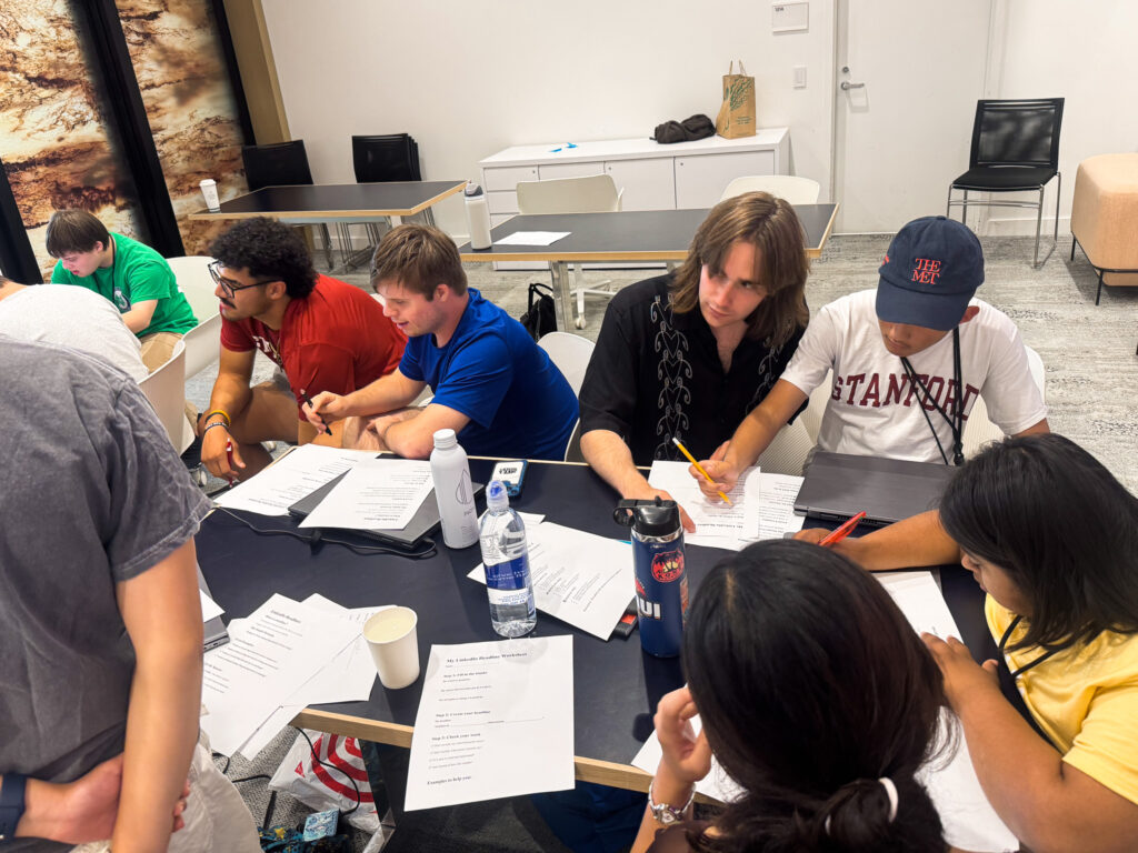 A group of intellectually or developmentally disabled youths work with Stanford students on a worksheet at a table in a conference room.