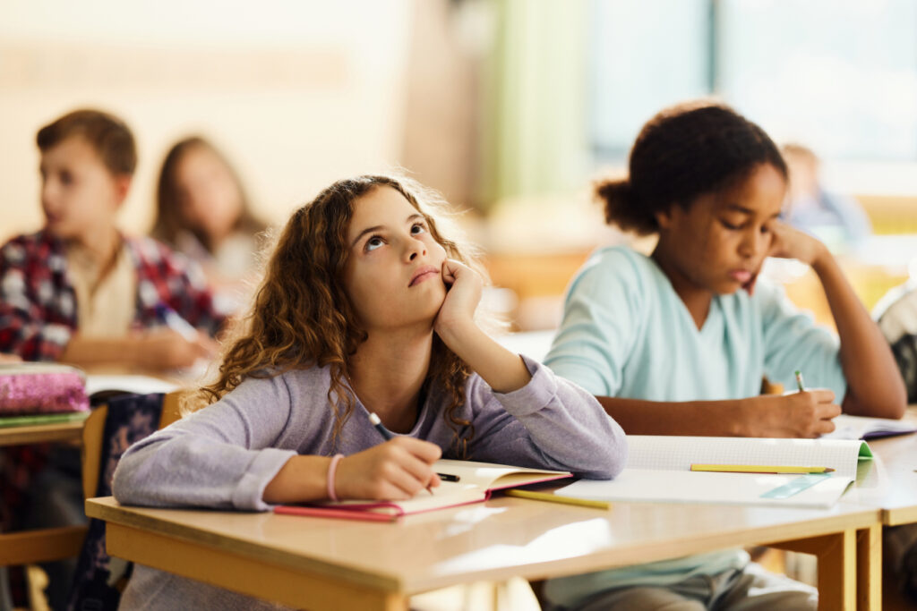 A bored-looking elementary school child sits at a desk in a classroom.