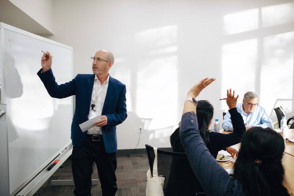A man writes on a whiteboard while people sitting around a table raise their hands.