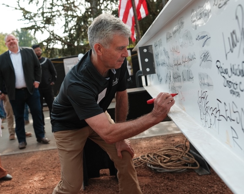 Dan Schwartz leans over while signing his name to a large white construction beam in front of a group of people.