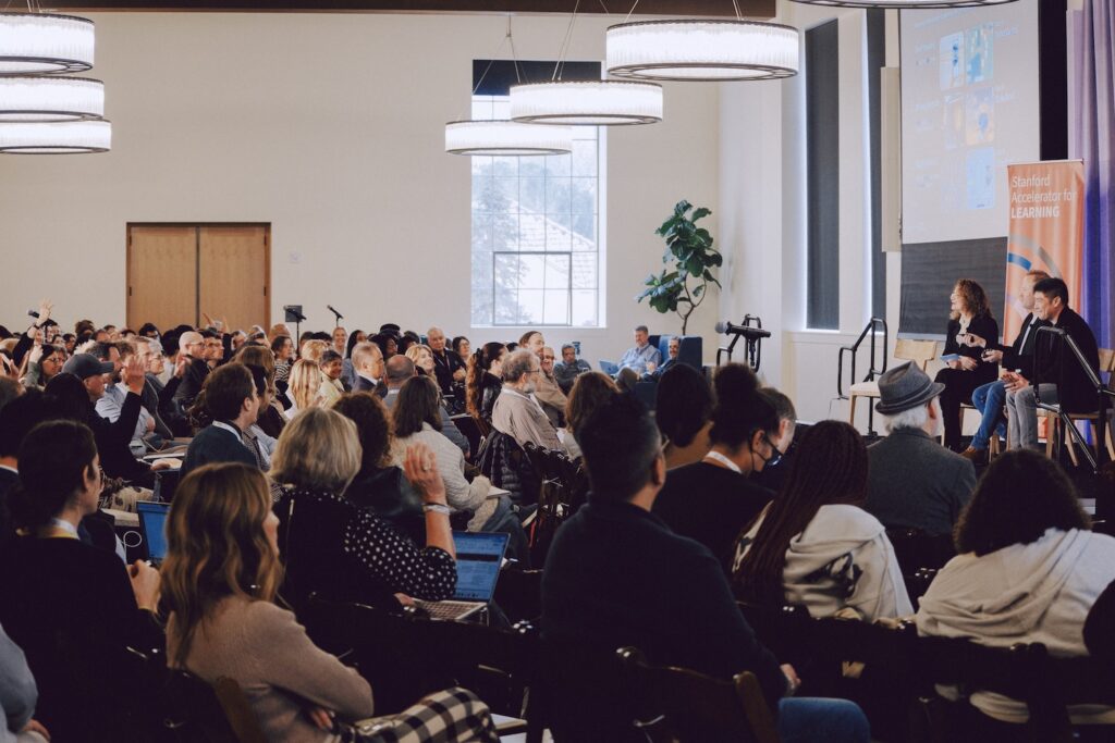A conference hall with beautiful natural light full of attendees and 3 presenters onstage.