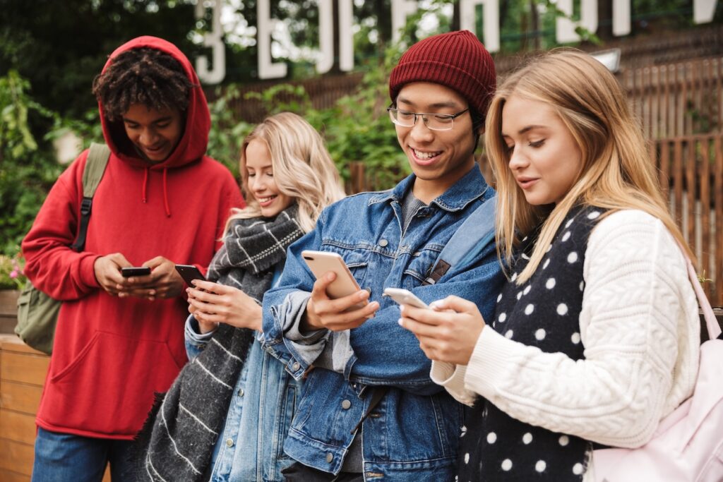 A group of teenagers cheerfully stand together while looking at their phones.