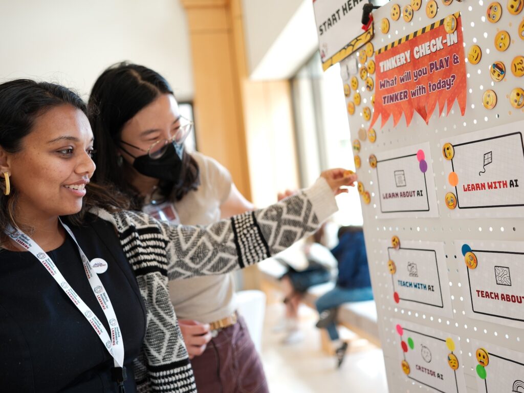 A south Asian woman and an east asian woman wearing a mask look at the AI tinkery posterboard together at an education summit.