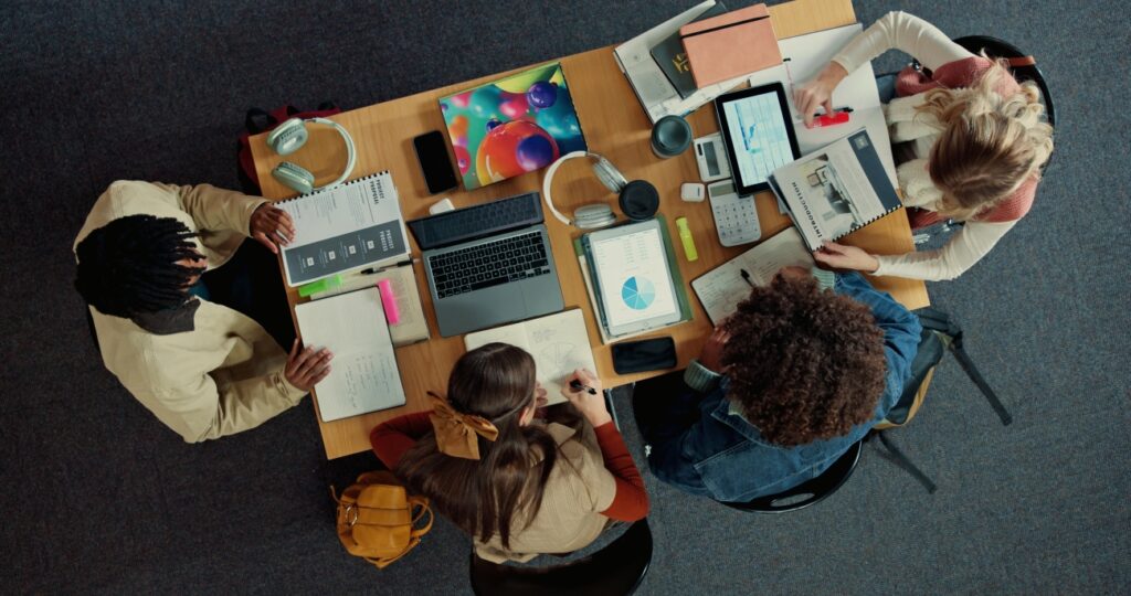 Top-down view of students studying at a table full of devices and notebooks.
