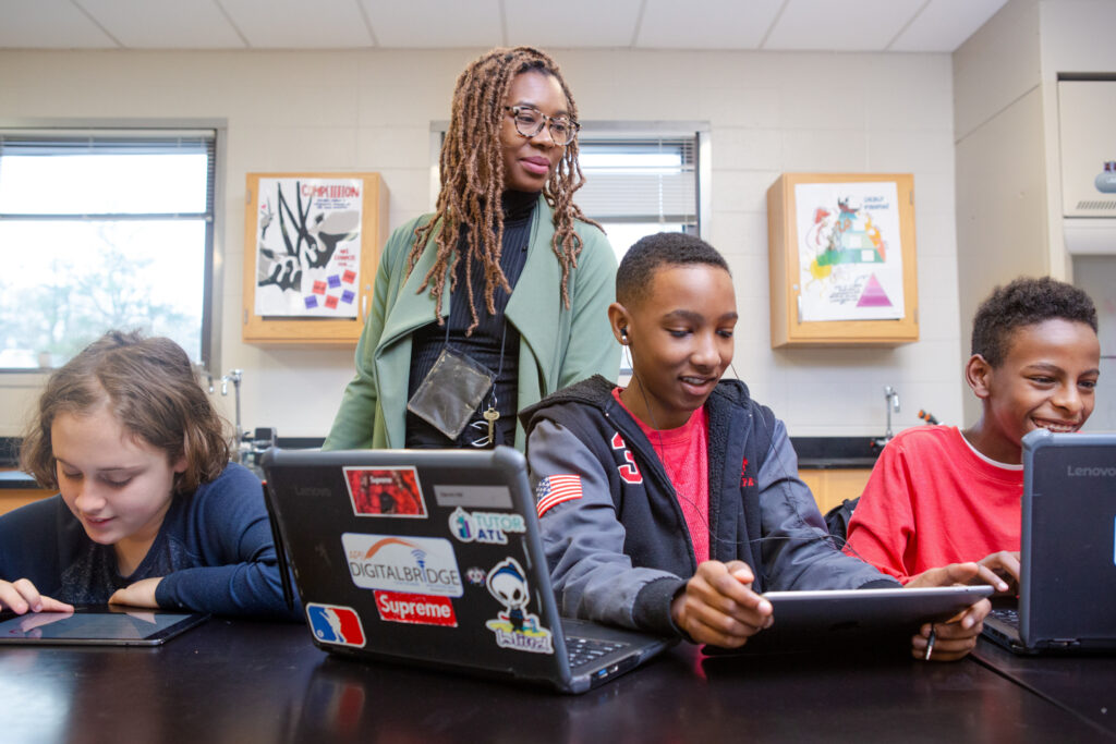 A teacher helps three boys working on tablets and laptops.