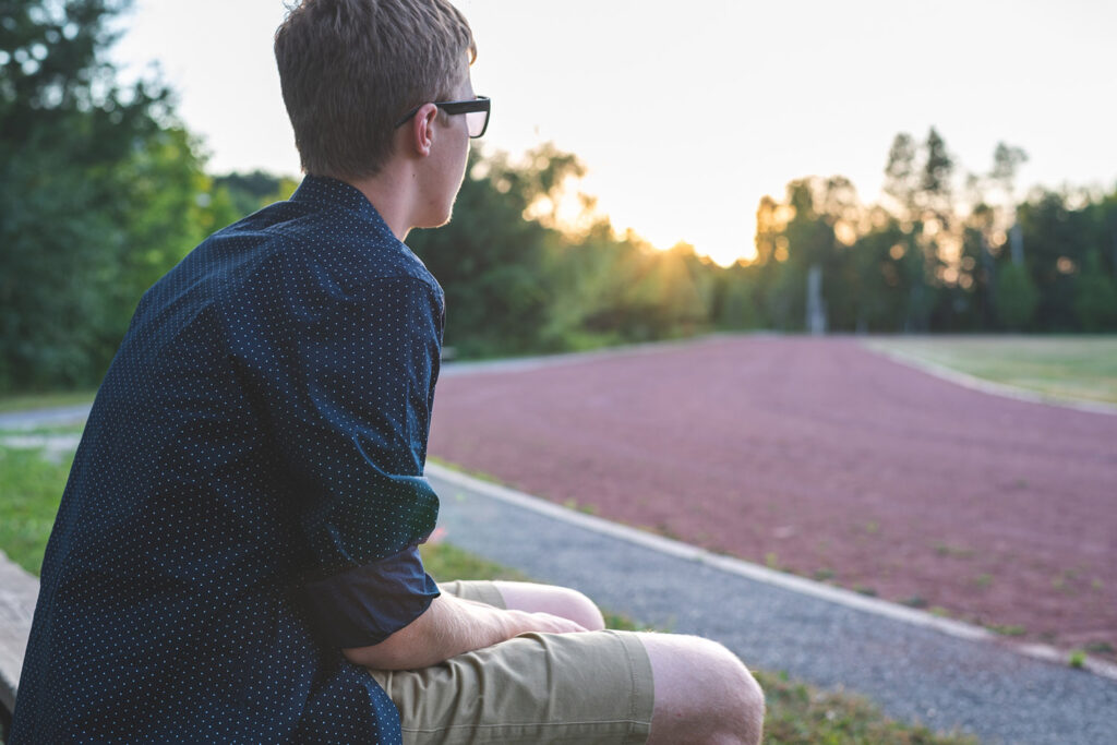 A young man sits on a bench looking out at a soccer field.